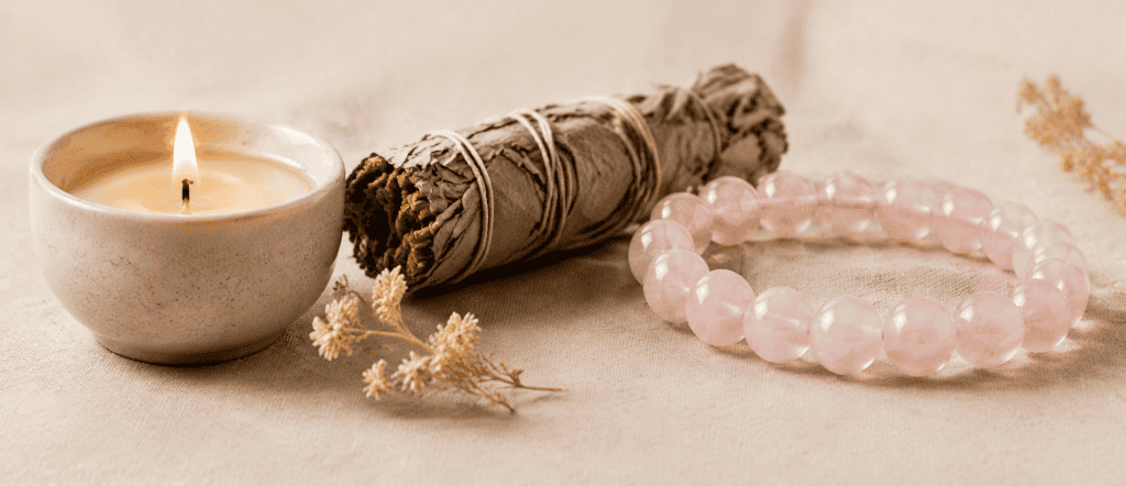 Soft wellness still life with lit candle, dried sage bundle, and rose quartz crystal bracelet on neutral fabric background